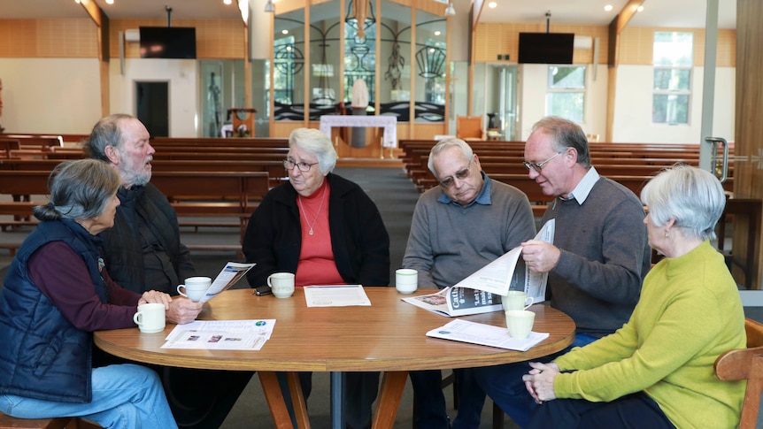 Six people sit at a table with newspapers and flyers and cups of tea