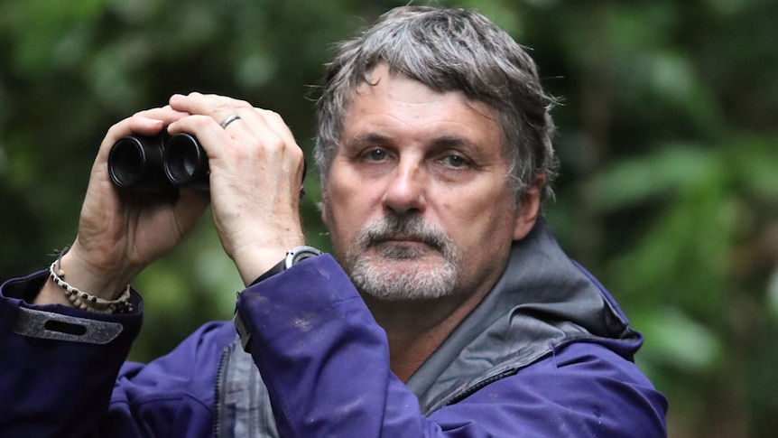 A man with grey hair and a beard holds up binoculars as he looks at the camera.