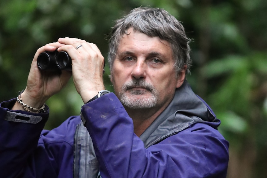 A man with grey hair and a beard holds up binoculars as he looks at the camera.