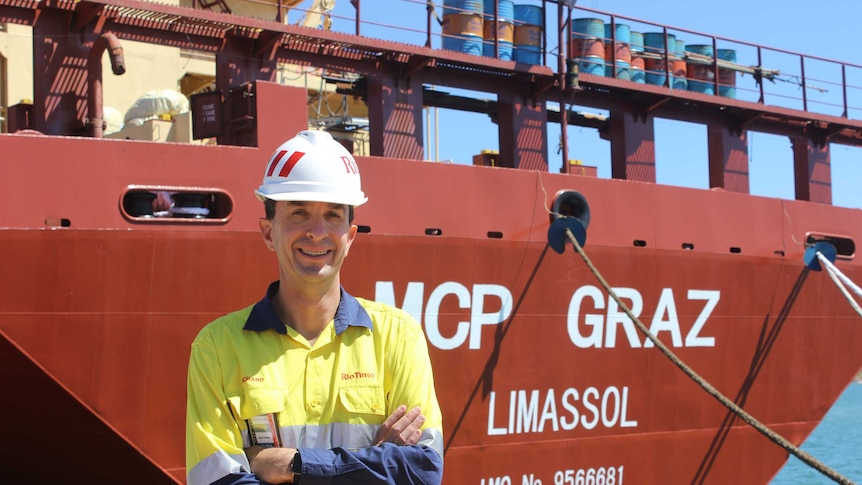 Man in long sleeved shirt and hard hat standing in front of a ship.