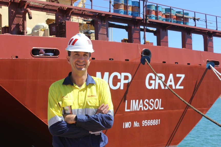 Man in long sleeved shirt and hard hat standing in front of a ship.