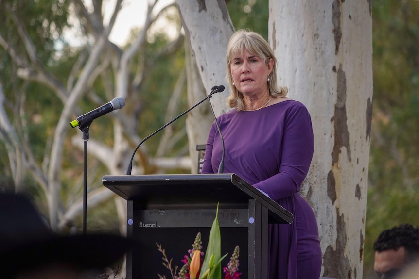 A middle aged woman wearing a purple dress standing at a lectern in a park full of gum trees, addressing the audience.