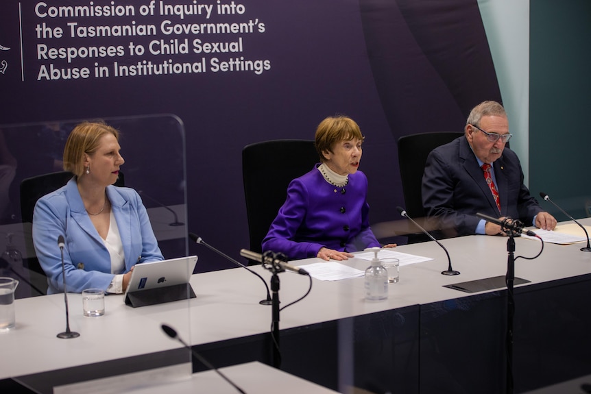 Two women and a man sit at a desk in front of microphones.