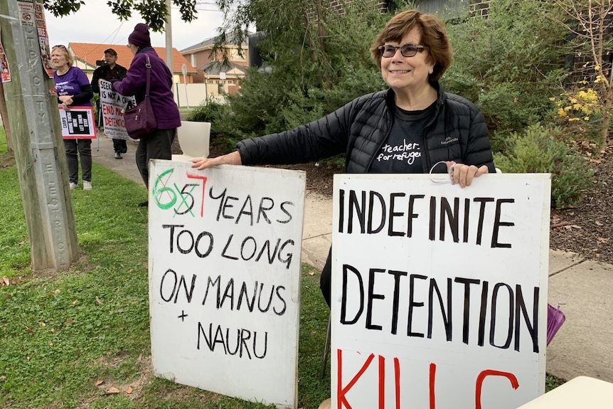 A woman in a black puffer jacket holds signs protesting indefinite detention on Nauru and Manus Island.