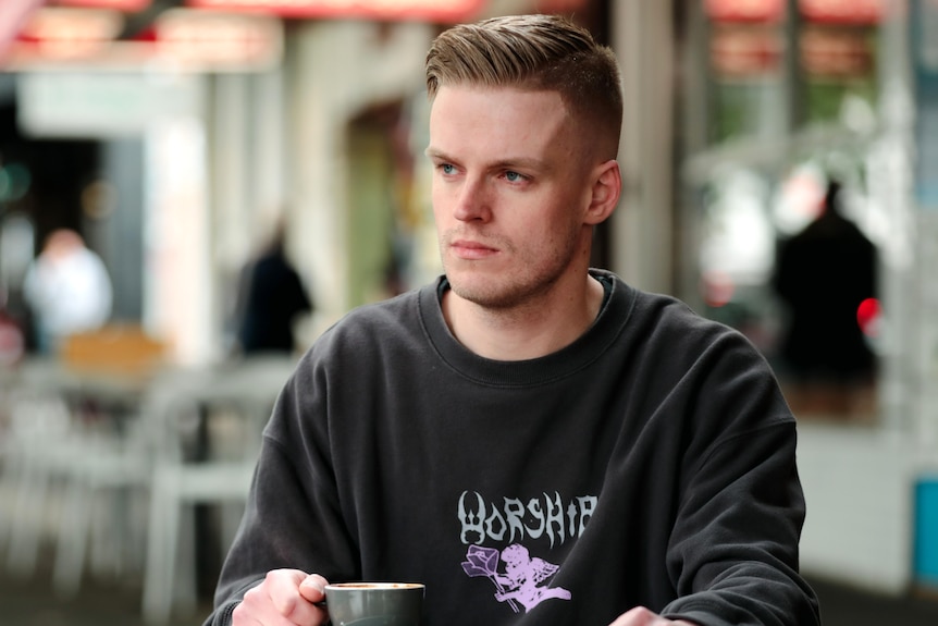 A young man sitting a cafe table, with a neutral expression, looking off to the side. 