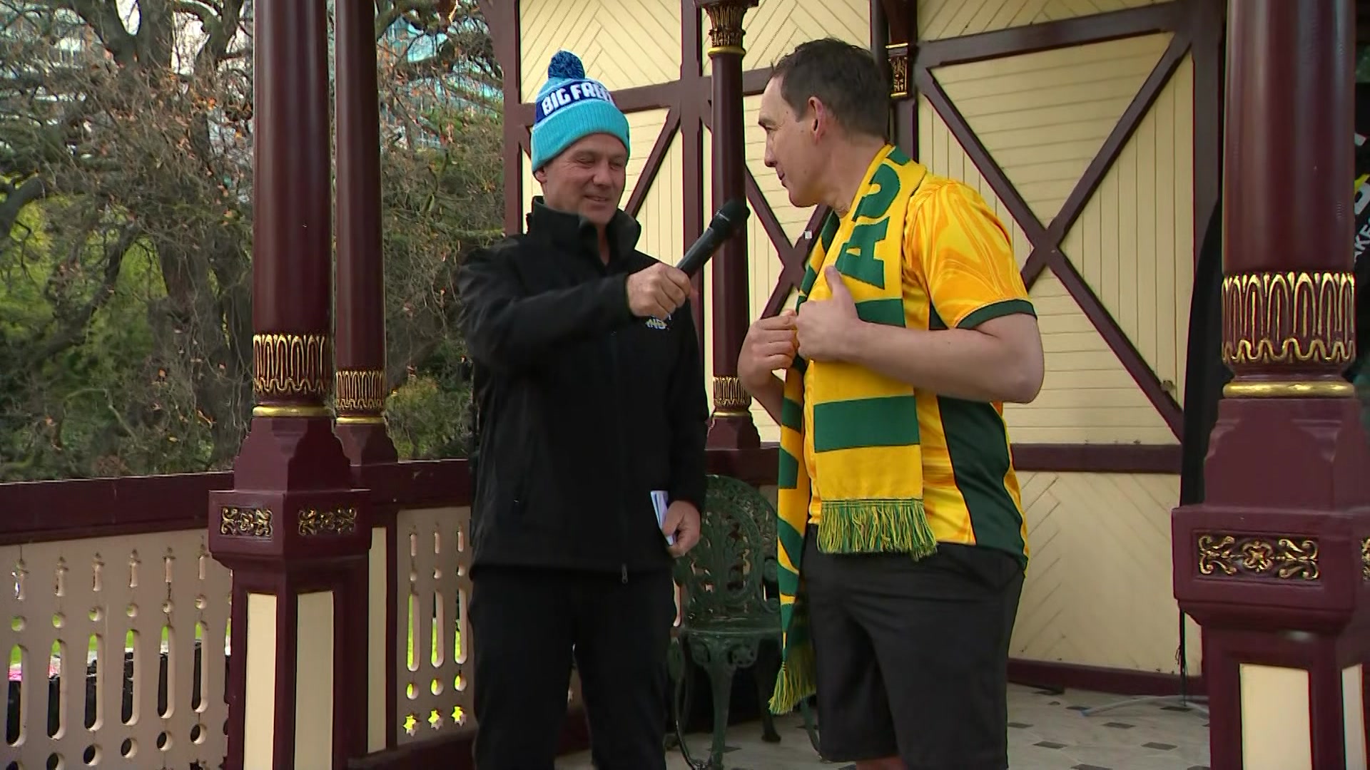A man in a blue beanie holds a microphone at a man wearing a yellow shirt, green shorts and green and gold scarf.