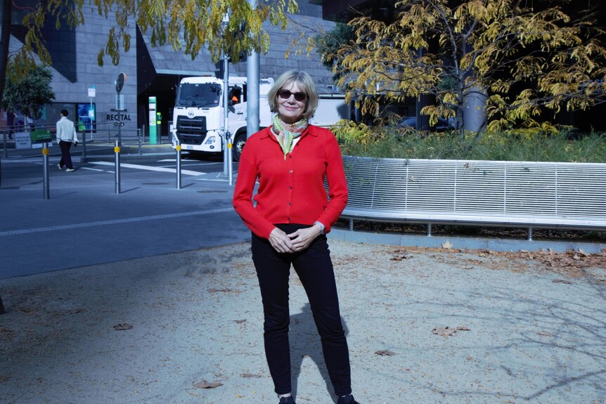 Kerstin Pilz, in black pants, red jumper and sunglasses, stands smiling with a busy city street behind her.