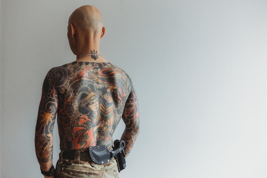 A bald shirtless man faces a white wall, his back is covered in elaborate tattoos