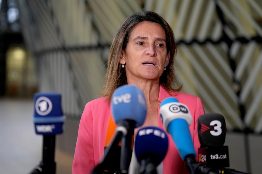 Una mujer con una chaqueta de traje rosa se para en medio de un discurso frente a cinco micrófonos.