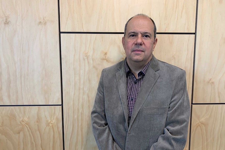 A man in a grey jacket stands in front of a wood-panelled wall.