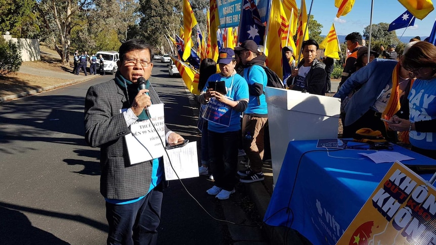 Chau Van Kham speaks at a protest in Canberra on 10 June 2018 with people holding yellow and red flags and signs.