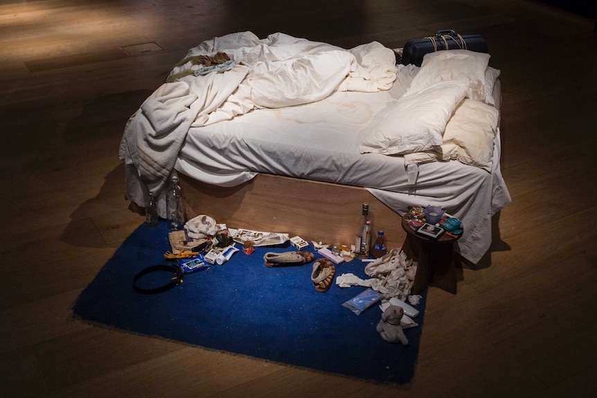 Colour photograph of artwork My Bed by Tracey Emin in a dimly lit gallery space.