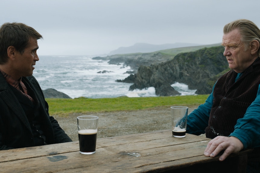 Two men sit at a wooden table with half-drunk glasses of stout in front of a spectacular Irish coastline backdrop.