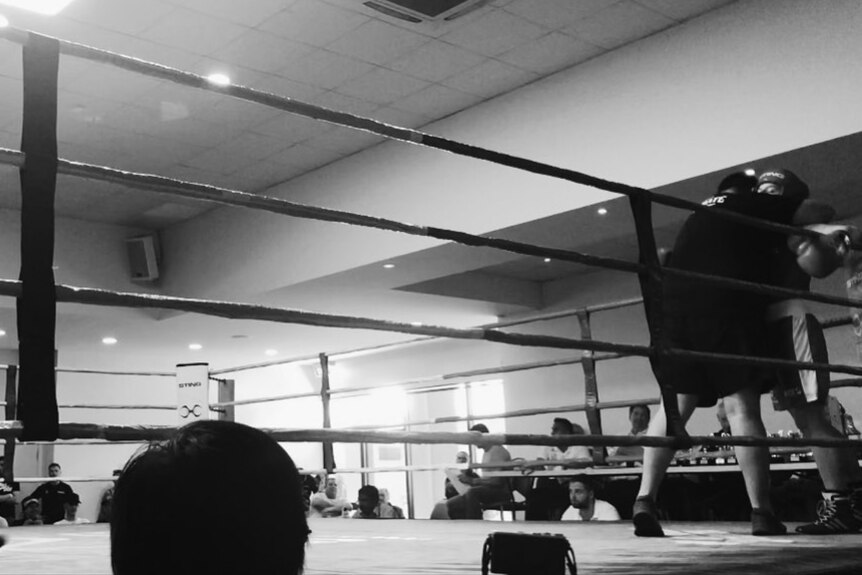 A blurry black and white photo of a ring, in a gym, with two fighters boxing infront of a crowd.