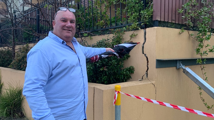 A man wearing a blue shirt points to a big crack in a retaining wall.