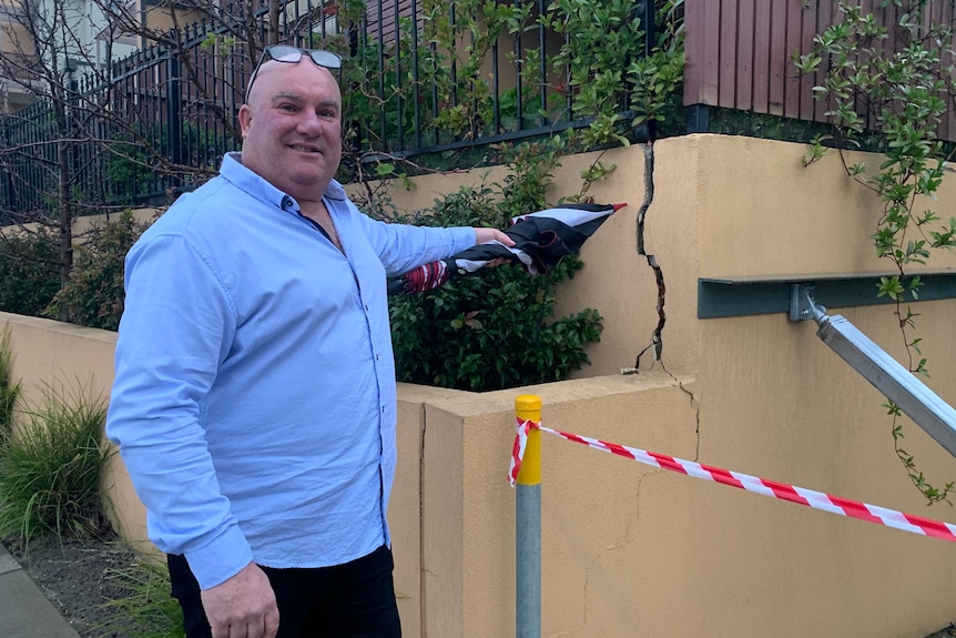 A man wearing a blue shirt points to a big crack in a retaining wall.