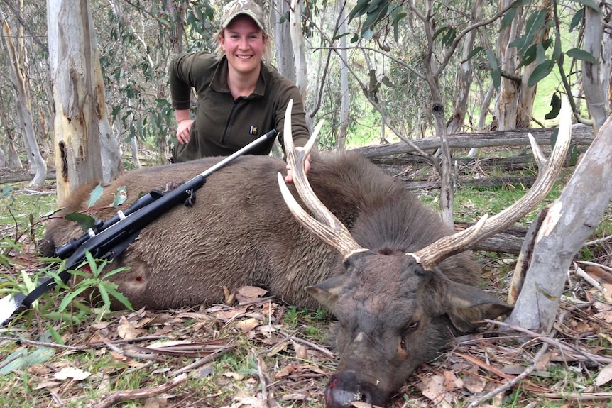 A woman kneeling behind a dead deer with her gun lying across its hindquarters.