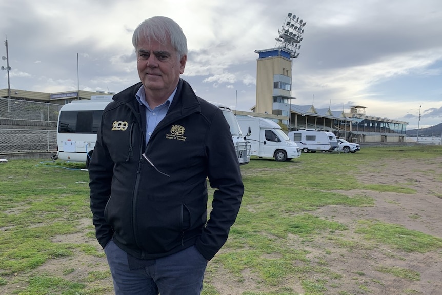 A man in blue pants and zip up jumper stands on showground with caravans behind him