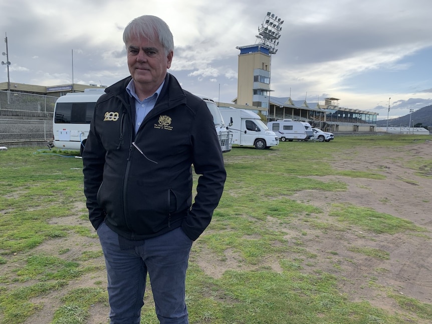 A man in blue pants and zip up jumper stands on showground with caravans behind him