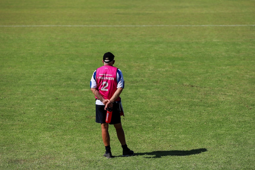 Older man standing wearing cap and pink high vis vest on green football oval.