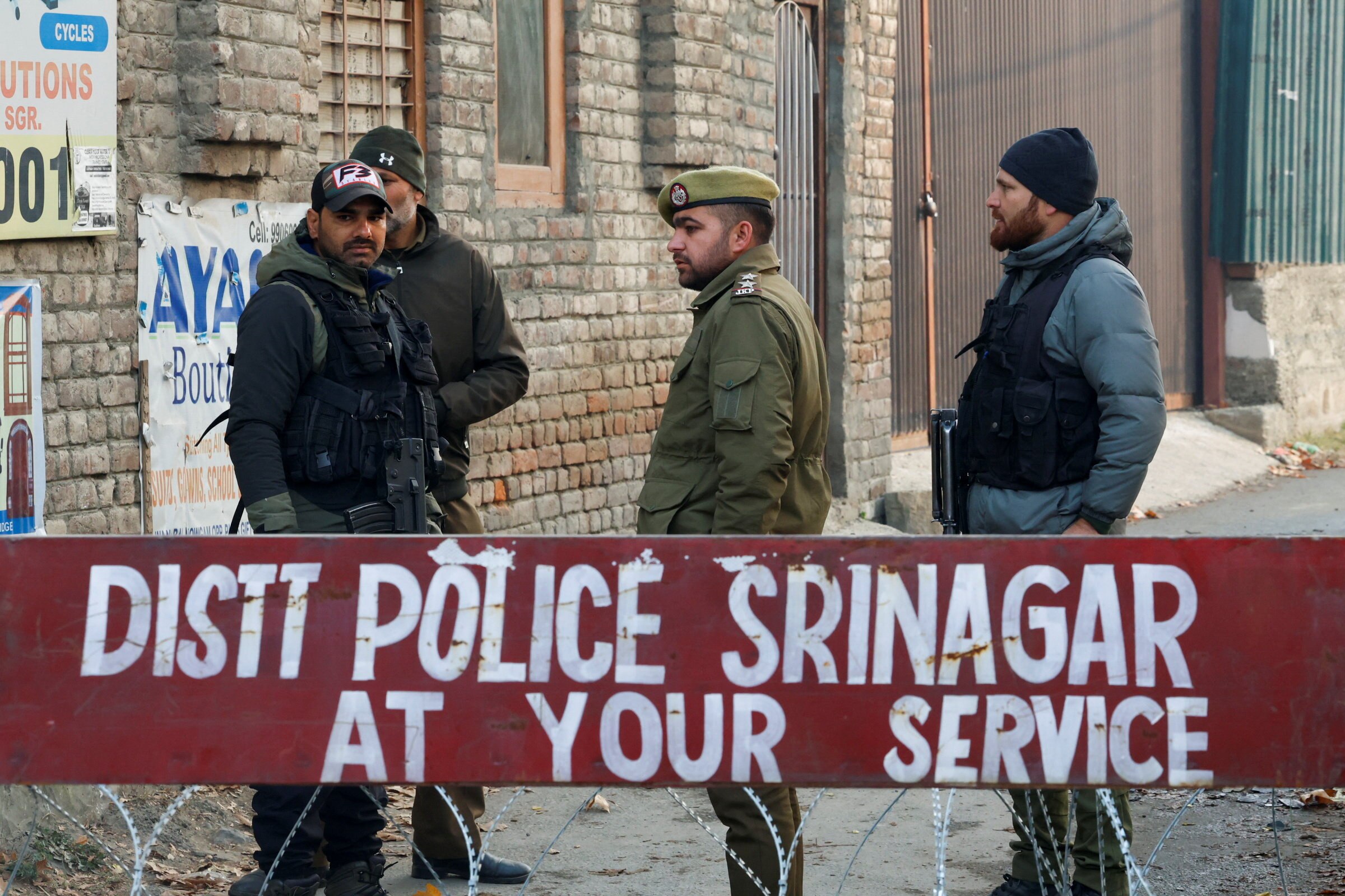 Four men, at least two carrying guns, stand behind a red sign reading 'Police Srinagar At Your Service'