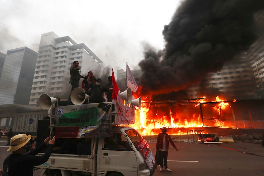 A truck carrying protesters and large megaphones drive down an empty thoroughfare as a metro station burns.