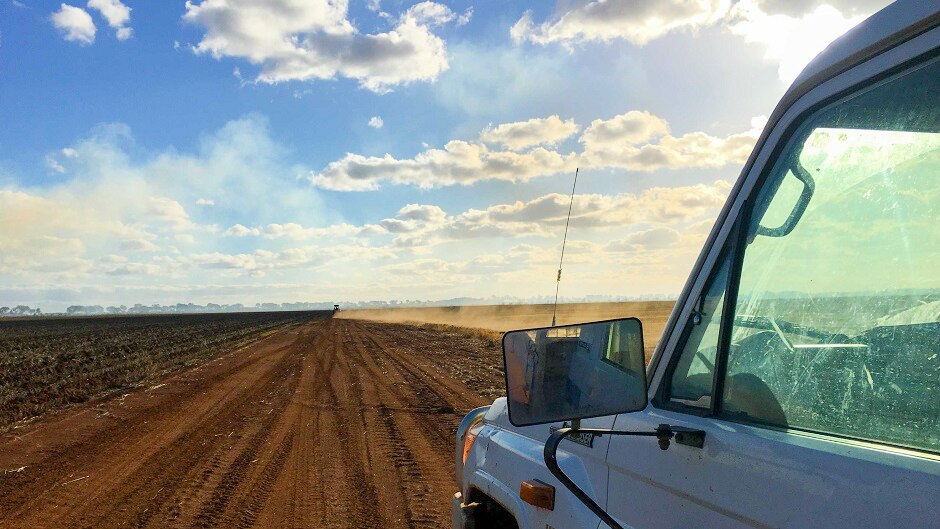 The passenger side of a ute in foreground with dust kicking up behind a vehicle on dirt road in the distance.