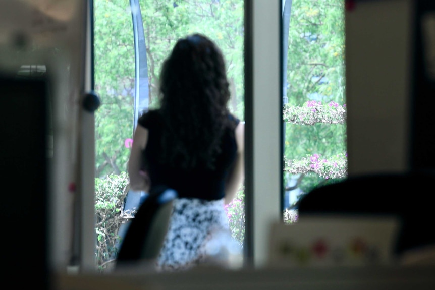 A woman looks out an office window at trees.