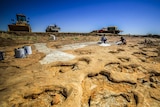 Wide divots in sandstone in the foreground, with people crouching in the dirt and bulldozers in the background under a blue sky.