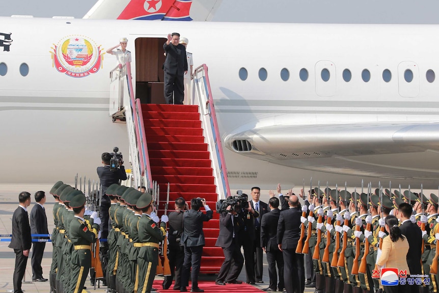 Kim Jong Un waves as he prepares to walk down steps covered in red carpet from a plane.