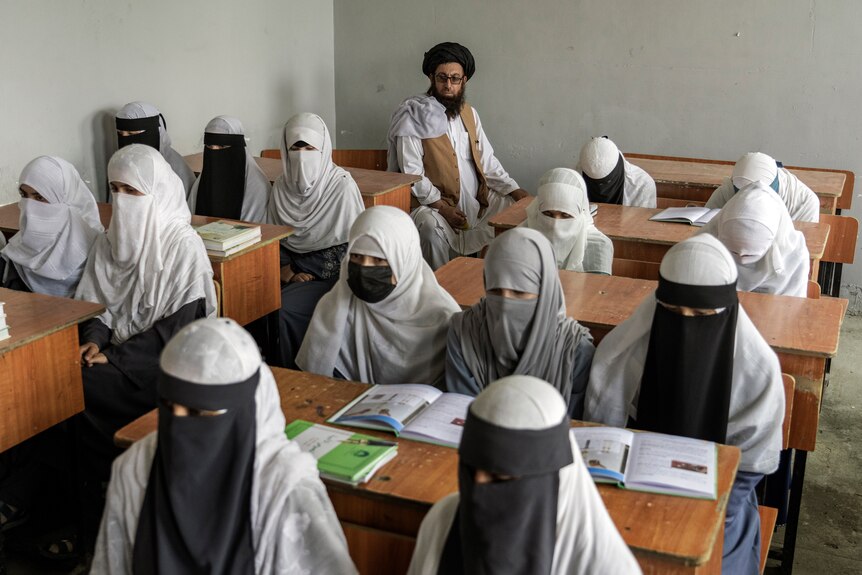 Girls in religious coverings sit at desks as a bearded man watches them from the back row. 