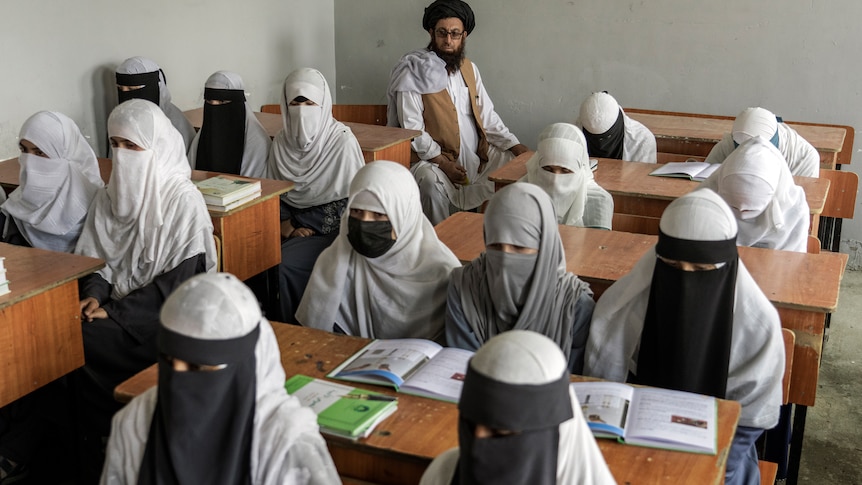 Girls in religious coverings sit at desks as a bearded man watches them from the back row. 