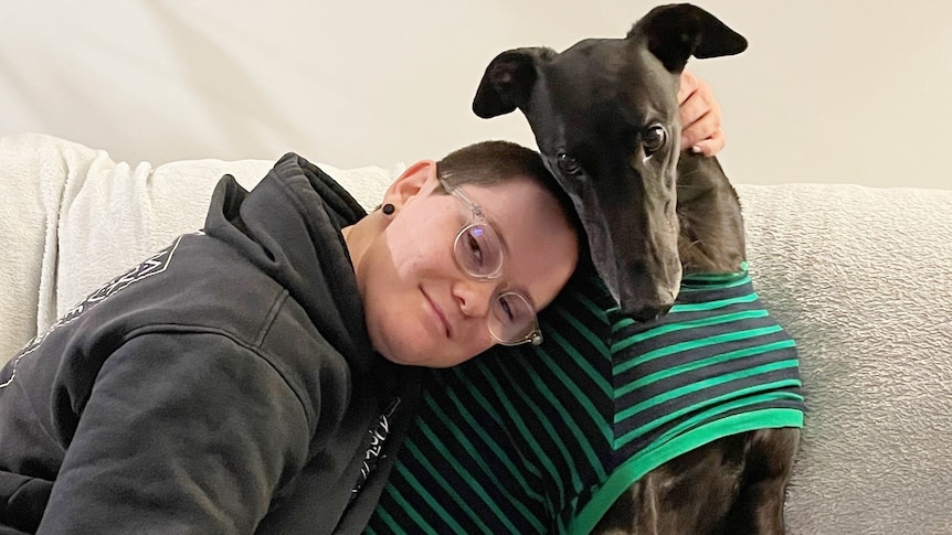 Women with glasses smiling at the camera while hugging a greyhound.