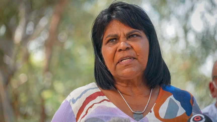 A medium close-up of a woman looking past the camera, speaking, in front of a bushland background.