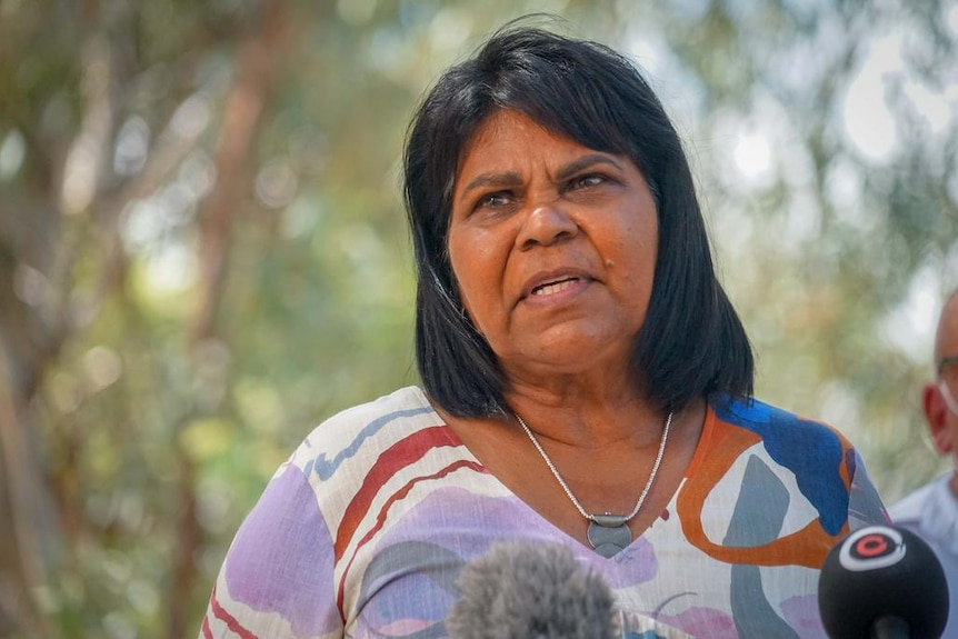 A medium close-up of a woman looking past the camera, speaking, in front of a bushland background.