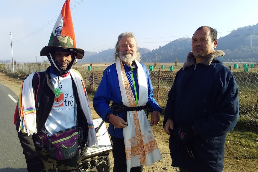 Three men standing in front of a fence and hill in India, with one wearing a cone-shaped Indian flag as a hat