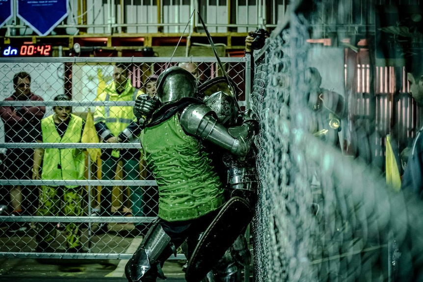 A fighter in armour kicks their opponent into the corner of the steel cage