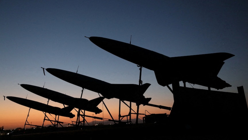 Aerial drones are silhouetted against a dimly lit sky. 