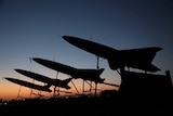 Aerial drones are silhouetted against a dimly lit sky. 