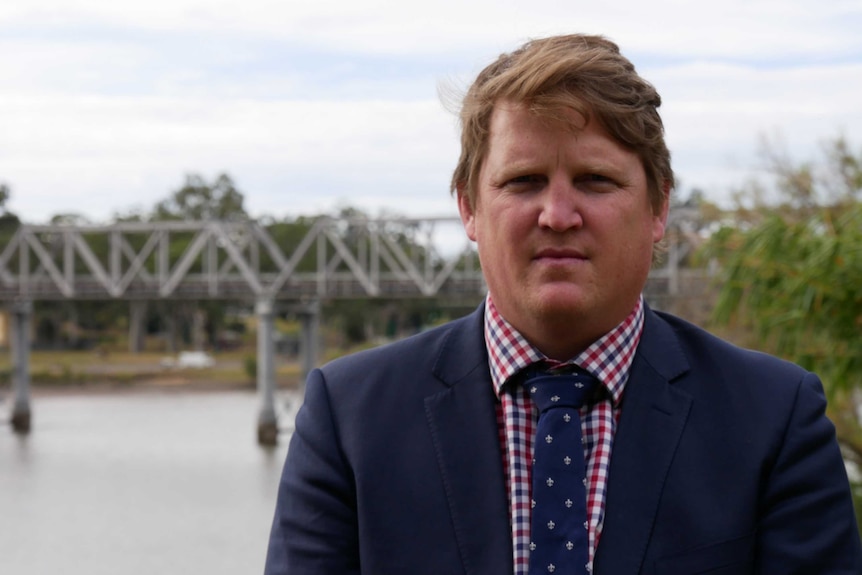 Middle aged man wearing a navy suit, red checked shirt and navy tie is standing in front of a river staring ahead, disappointed