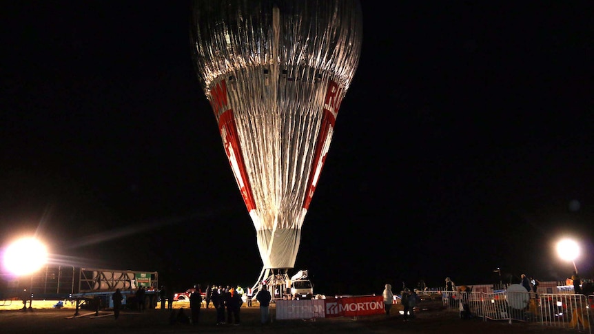 A tall silver hot-air balloon stands in darkness in a field, with people on the ground below.