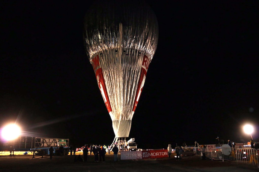 A tall silver hot-air balloon stands in darkness in a field, with people on the ground below.