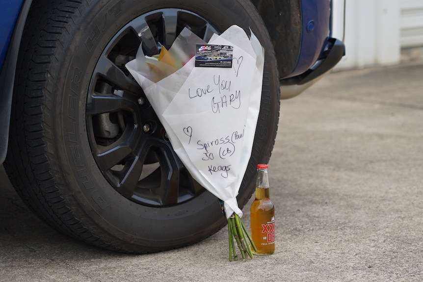 A bouquet of flowers reading "Love you Gary" and a beer leaning up against a car wheel.
