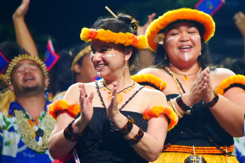 Performers from Guam wearing orange flower headbands clap, smiling.