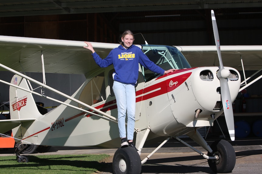 A smiling young girl stands on the wing of a light aircraft with her arms on the plane