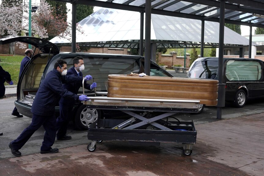 Two men in navy suits wheel a wooden coffin away from a black hearse parked in a line with others.