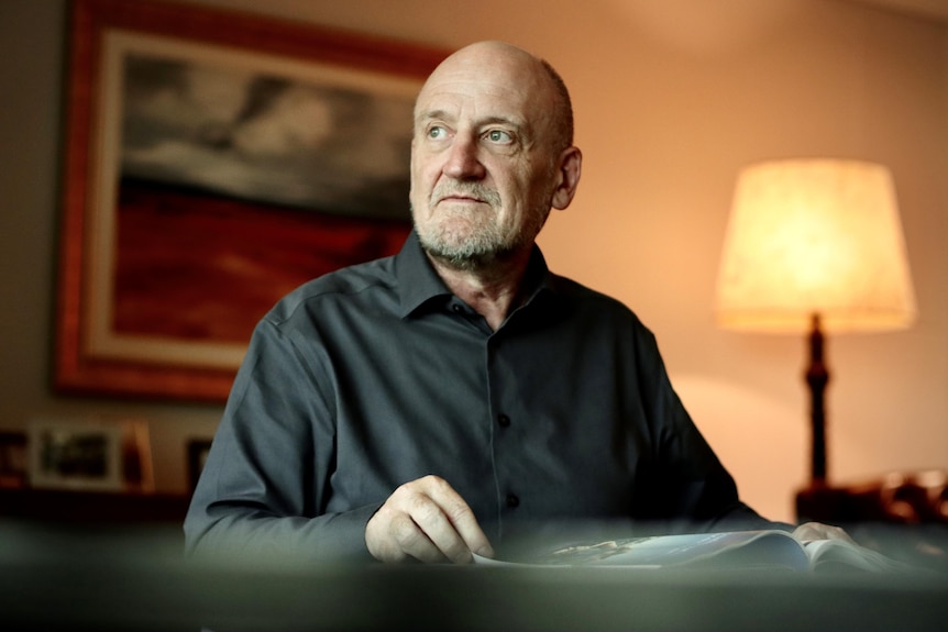 An older man wearing a dark shirt sits behind a desk and looks out to the side of the picture.