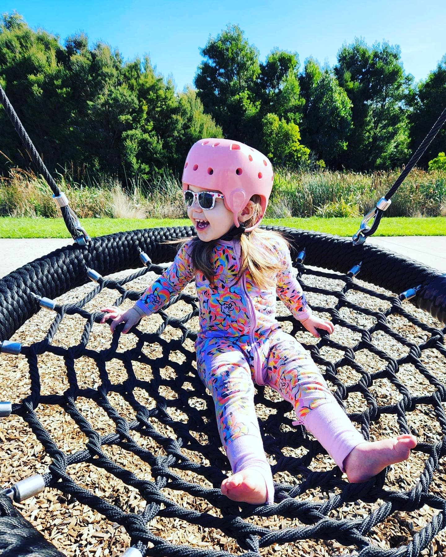 A child playing on a circular netted swing at the park, wearing a pink helmet as well as sunglasses.