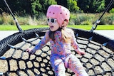 A child playing on a circular netted swing at the park, wearing a pink helmet as well as sunglasses.