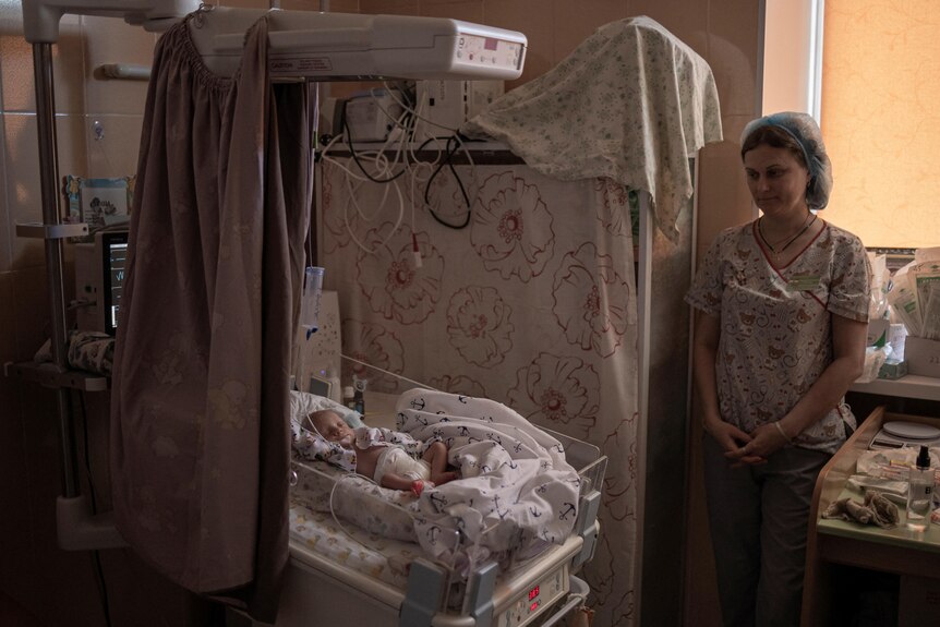 A nurse stands with her hands clasp next to a baby in a hospital crib. 